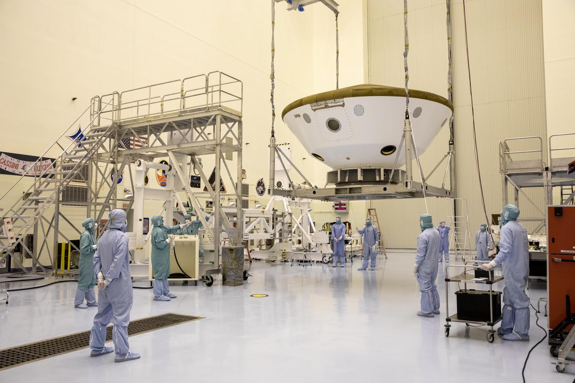 Several workers wearing white and blue protective suits watch as the Mars spacecraft is moved with a crane on a metallic lift at NASA's Kennedy Space Center in Florida in August 2024. Photo credit: NASA/Kim Shiflett